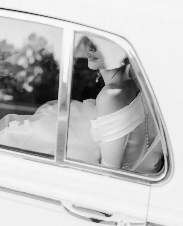 Noiva dentro de um carro, vista pela janela, com vestido e véu brancos