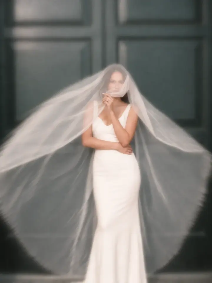 Noiva de vestido branco sob um véu longo, diante de uma porta escura